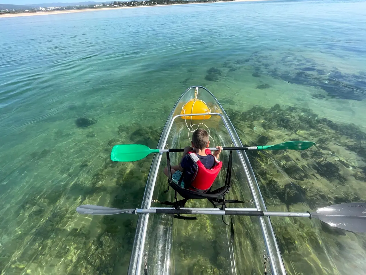 A child paddling a clear kayak, seen from behind, with seagrass visible through the hull